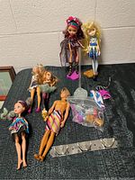 Five dolls including four female dolls and one male doll displayed on a table along with accessories and stands.