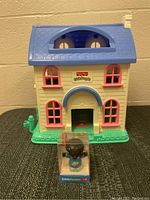 Front view of the Fisher Price Little People toy house with distinctive blue roof, pink window frames, and green base on the floor.