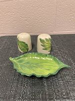 Pair of ceramic salt and pepper shakers with green leaf decoration and matching green leaf-shaped dish on a dark textured surface.