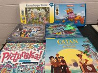 Photo showing 6 games and puzzles stacked including Ravensburger puzzle, Big Potion, Pictopia, Catan Junior, Pictureka, and Jeopardy.