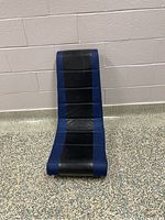 Front view of blue and black gaming floor chair seated against a white wall on a speckled floor.
