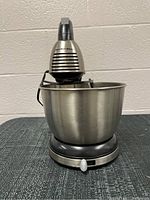 Front side view of Hamilton Beach stand mixer with stainless steel bowl on dark textured surface against white wall.