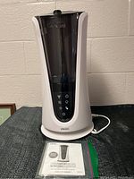 Front view of the white and black Homedics humidifier with control panel and manual in front on fabric surface. Shows overall condition and included manual.