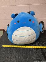 Front view of the large blue Squishmallow showing face, horns, belly pattern, and measurement tape for size.