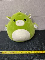 Front view of green dragon Squishmallow plush showing its face, belly patch, ears, and spikes with a measuring tape showing about 12 inch height.