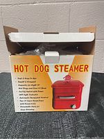 Boxed hot dog steamer showing front and side with features listed including capacity, thermostat, LED indicator, and detachable parts.