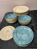 Set of 8 large cream dinner plates stacked behind 7 blue patterned bowls stacked, one blue bowl facing up in front, and 8 cream plates stacked with one turned over showing Pfaltzgraff mark.