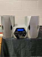 Front view of Scott radio/CD player flanked by two matching silver speakers on a black cloth. The radio display is blue-lit.