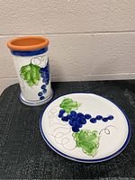 Full view of wine bottle cooler alongside matching plate on a black cloth surface showing grapevine painted design with blue grapes and green leaves.