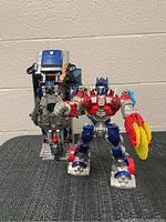 Two Transformers Optimus Prime figures, one with a blue cab unit and mechanical lower body, the other with red, grey, and blue robot body holding a yellow saw blade on its right arm.