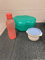 Photo of pink translucent water bottle, large green segmented bowl, and small white bowl with blue lid placed on a dark surface against a beige wall.