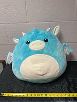 Front view of turquoise blue and white Squishmallow plush dragon with tape measure for size reference showing about 15 inches width.
