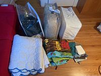 Photo of multiple folded bed linens and bedding items arranged on floor beside red couch, including white quilted comforters, Winnie The Pooh sleeping bag, and bedding stored in plastic bags.