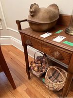 Full view of the chicken-shaped clay roaster on wooden furniture with two small woven baskets below, one filled with dried flowers.