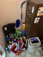Photo shows an assortment of various household cleaning supplies including spray bottles, bleach container, brushes, duster, dustpan, bucket, and plunger near filing cabinet and wall.