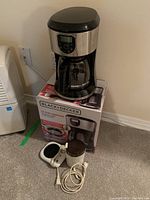 Photo showing the Black+Decker coffee maker on its original box, alongside the electric coffee grinder and white mug heating pad with power cord, placed on carpeted floor.