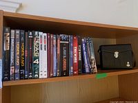Photo of a wooden shelf containing multiple DVD cases of TV series including Breaking Bad, Homeland, Law & Order, Big Bang Theory, JAG, The Wire, and Parks and Recreation, alongside a closed black case for binoculars.
