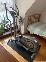 Full view of Tunturi elliptical machine standing next to bed on black rubber mat.