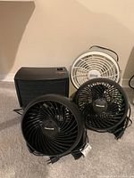 Four small home electric appliances: two black Honeywell fans, one black ThemaFlo heater/fan, one white Whirlwind fan on beige carpet.