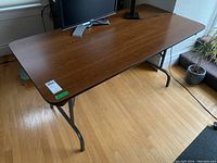 Rectangular folding table with brown laminated particle board top and black metal folding legs, viewed from above corner angle against wooden floor and window.