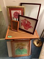 Overall photo showing multiple framed items including needlework, paper cutting, blank frame and drawing stacked on a wooden cart and leaning on the wall
