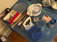 Overview of assorted kitchenware items arranged on blue tablecloth, showing rolling pins, food storage containers, bagel cutter, grater, cooking utensils, and coasters.