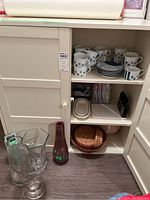 Mikasa cups with matching saucers, wicker baskets, glassware including infusing teapot and mugs on white shelves.