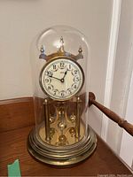 Full view of Kundo anniversary clock under glass dome showing clock face, brass base, and pendulum