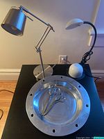 Two metal desk lamps, metal bowl, and two scissors shown on dark table against wall.