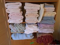 Shelf filled with neatly folded bathroom towels and facecloths in cream, beige, blue pastels, with folded bed linens in white, pastel purple, and printed fabrics below. Thick-knit cream blanket on the bottom shelf.