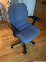 Blue upholstered office chair with black armrests and five-wheel black base on wood floor in front of closed white door and wooden cabinet.