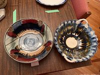 Top view of two pottery bowls placed on a wooden table, showing one round bowl with multicolor glaze and one scalloped bowl with handles and blue/brown glaze.
