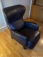 Front angle view of black bonded leather recliner chair showing padded backrest, seat and armrests, with wood legs on hardwood floor near white wall and closet doors.