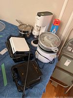 Photo showing assorted kitchen appliances on a blue tablecloth including sandwich makers, double boiler, and rice cooker.
