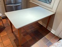 White rectangular kitchen table with natural wood legs standing on a tile floor near a window and door showing light wear on the top surface.