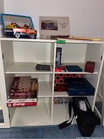 White cabinet containing board game boxes Scrabble and Stratego, Playmobil toy trucks on top shelf, and cases and boxes including Trivial Pursuit and other games