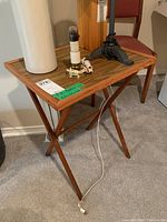 Photo showing one wooden TV tray table with a rectangular top and folding legs, holding a lamp and other household items. The table has some scuffs and is set on carpet.