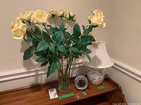 Photo showing a clear glass vase with cream faux roses, metal and plastic alarm clock, and ceramic base desk lamp on a wooden surface.