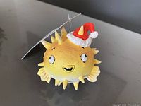 Yellow puffer fish shaped Christmas ornament with large eyes and a Santa hat, photographed against a neutral background.
