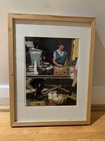 Framed color photograph featuring a woman behind a market stall with vegetables and a scale, in a light wood frame with white matting.