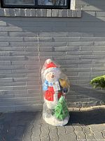 Photo of the snowman figure wrapped in plastic against a light gray brick wall, showing full figure with red hat, scarf and Christmas tree.