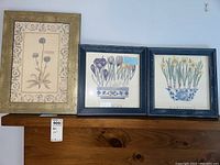Three framed floral botanical prints displayed on wooden furniture to show the full set and frame styles.