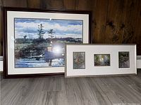 Two framed pictures positioned on floor against wall. One large single painting of fishing scene, one framed triptych of landscapes.
