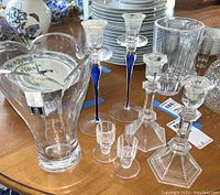 Photo showing three pairs of clear glass candlesticks including two tall with blue stems, two shorter hexagonal base, and two very short ones, placed near dishes on a wooden table.