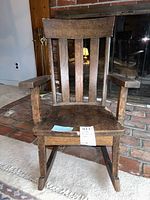 Front view of a child's wooden rocking chair showing vertical slats on the backrest, solid wooden seat, and flat armrests, with visible scratches and wear.