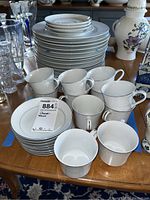 Photo showing stacked dinner plates, smaller plates, tea cups, and saucers on a wooden table with other items.