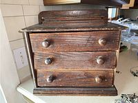 Front view of a wooden three-drawer jewelry box showing wear and surface scratches; the drawers have round wooden knobs and appear slightly misaligned.