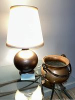 Metal table lamp with round base and white lampshade beside a footed copper bowl with handle on glass table, both showing some surface wear and scratches.