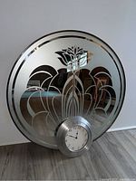 Round wall mirror with floral engraved pattern and integrated silver frame clock at bottom center, placed on floor against wall.