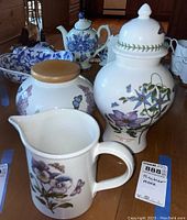 Photo showing pitcher, jar with wooden lid, and tall lidded vase with botanical prints on wooden table.